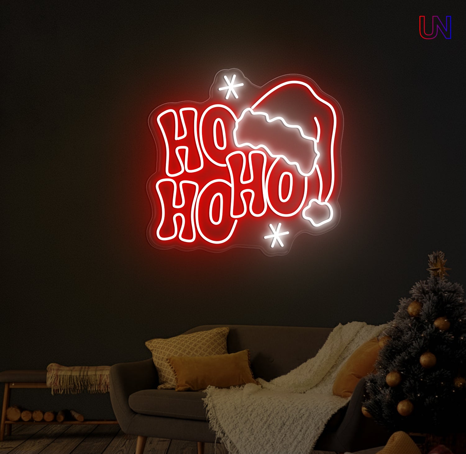 Red and white "Ho Ho Ho" text with a Santa hat and stars, illuminated as an LED neon sign on a dark wall above a grey sofa with a Christmas tree to the right.